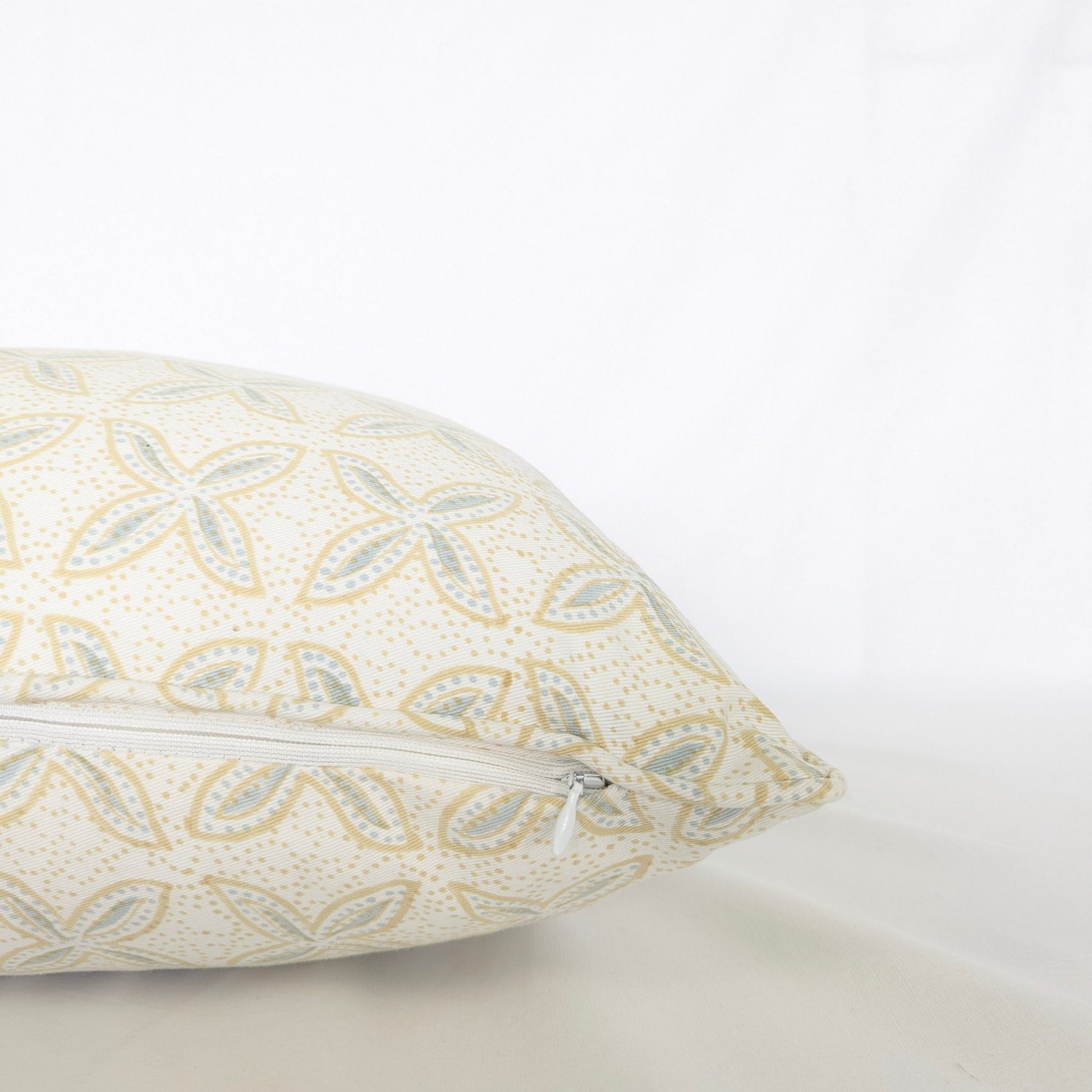 A close-up side view of the hand-block printed Bali plumeria Breeze cushion resting on a crisp white background. This detailed shot highlights the neatly sewn discreet hidden zipper closure. The soft blue and tan floral geometric pattern showcases the quality of this handcrafted textile, perfectly adding a light, cozy modern rustic Canadian farmhouse aesthetic to your home decor.