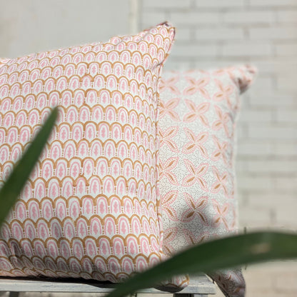 Close-up of two 100% cotton block printed pillow and cushion covers. The front cover features a pink and tan scalloped arch pattern, while the rear cover displays a soft pink floral cross design on white fabric. Partial view of a green plant in the foreground.