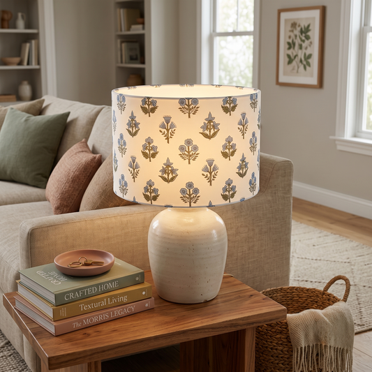 A warm glowing table lamp featuring a white ceramic base and a delicate light blue and green floral sprig drum lampshade. Resting on a wooden side table beside a neutral sofa, this piece perfectly adds a cozy modern rustic farmhouse aesthetic to Canadian and USA living rooms.