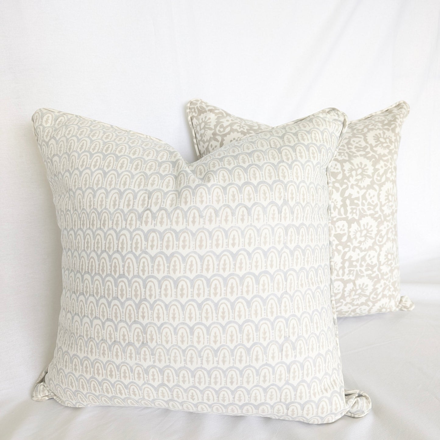 Two beautifully styled hand-block printed cushion covers resting on a crisp white surface. The foreground pillow showcases a pale blue and neutral beige scalloped arch pattern, layered perfectly in front of a complementary beige floral motif pillow. These artisanal square throw pillows are ideal for adding a warm, sophisticated modern rustic farmhouse aesthetic to both Canadian and USA homes.
