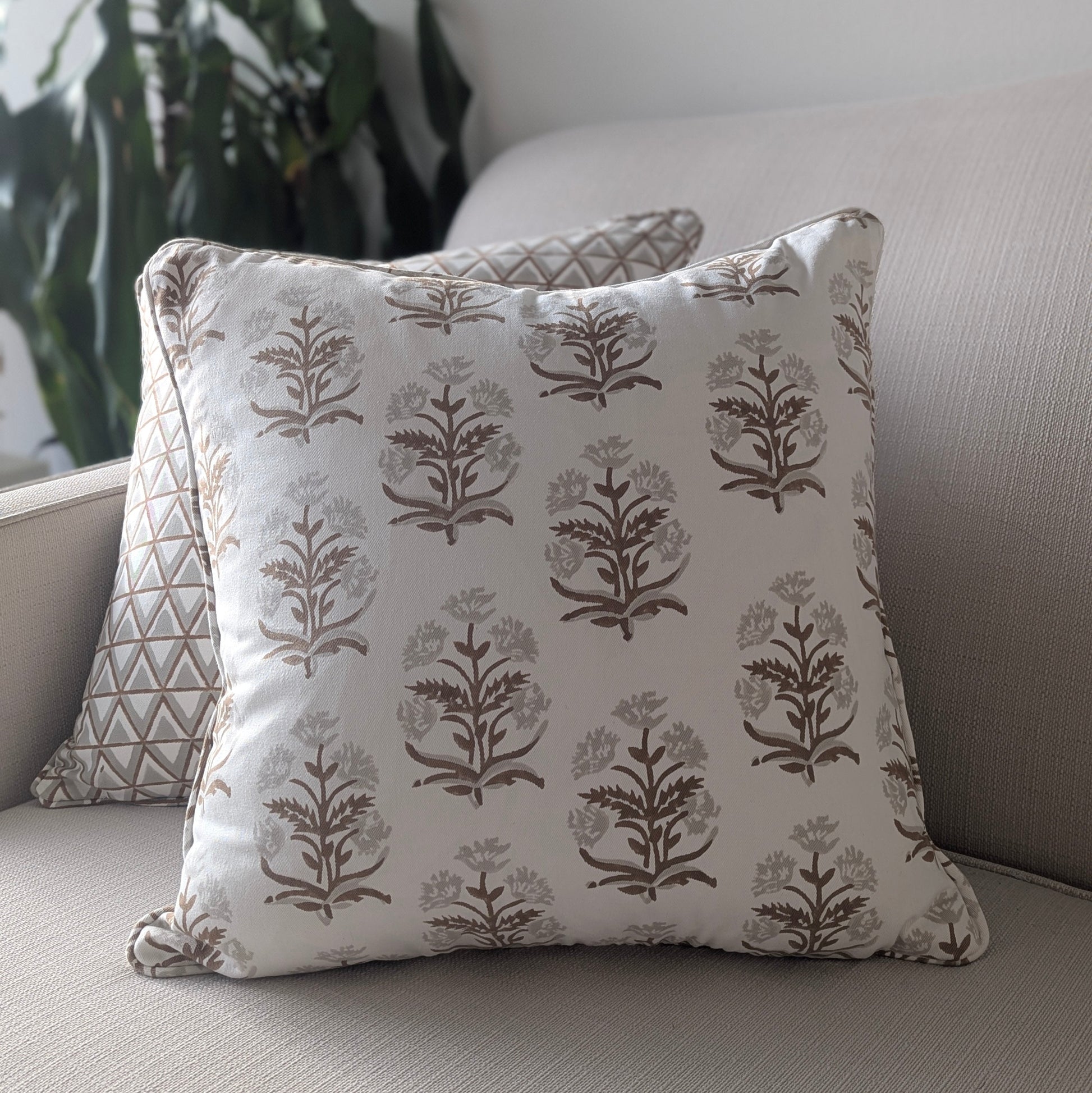 A close-up of a hand-block printed square cushion featuring an earthy brown and taupe floral plant motif on a cream background, resting on a neutral textured sofa. Behind it sits a partially visible coordinating geometric triangle patterned pillow. These beautifully handcrafted textiles perfectly add a cozy modern rustic Canadian farmhouse aesthetic to your living room decor.