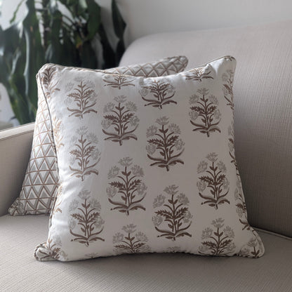 A close-up of a hand-block printed square cushion featuring an earthy brown and taupe floral plant motif on a cream background, resting on a neutral textured sofa. Behind it sits a partially visible coordinating geometric triangle patterned pillow. These beautifully handcrafted textiles perfectly add a cozy modern rustic Canadian farmhouse aesthetic to your living room decor.