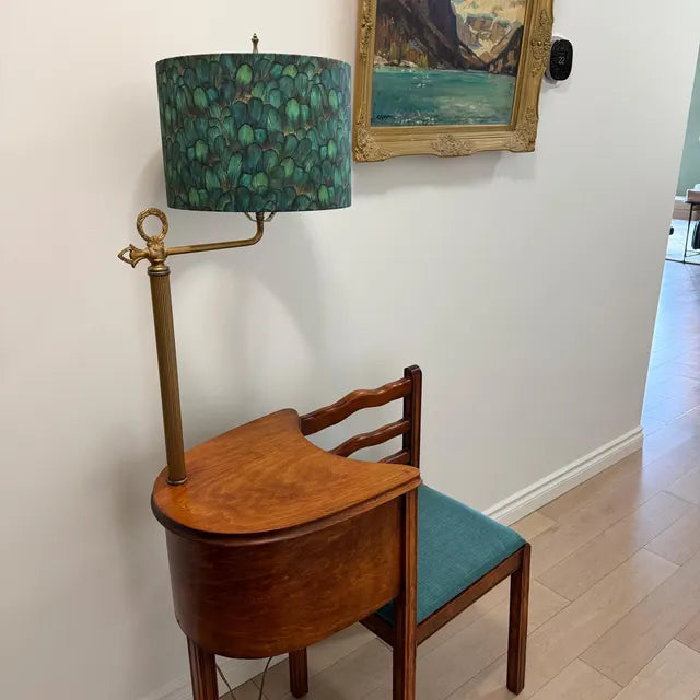 A vintage-style wooden writing desk with a curved front and a brass lamp attached to it, paired with a matching wooden chair that has a teal seat cushion. The setup is placed against a white wall in a home interior next to a framed landscape painting.