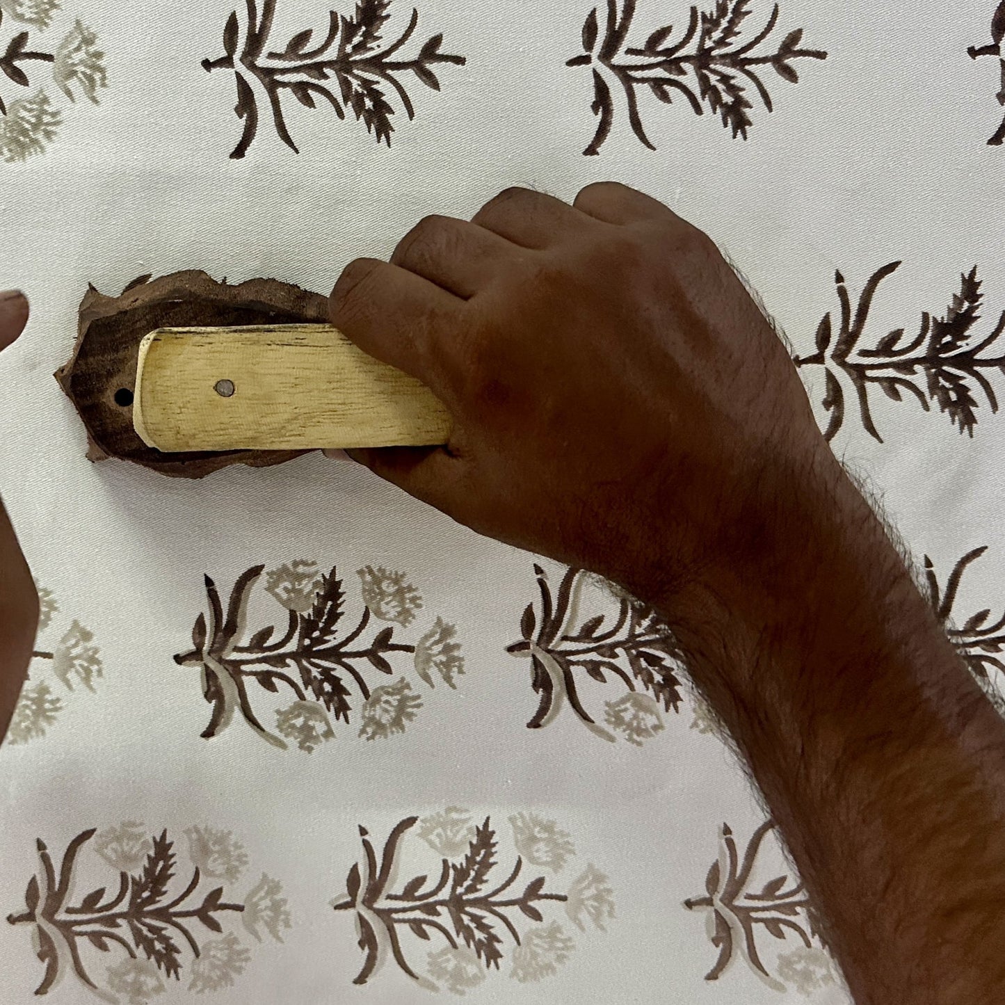 A close-up, top-down view of an artisan's hand pressing a carved wooden block onto cream fabric, stamping an earthy tan and brown botanical floral motif. This showcases the traditional hand-block printing process for handcrafted textiles, highlighting the authentic, artisanal quality that perfectly adds a cozy modern rustic Canadian farmhouse aesthetic to your home decor.