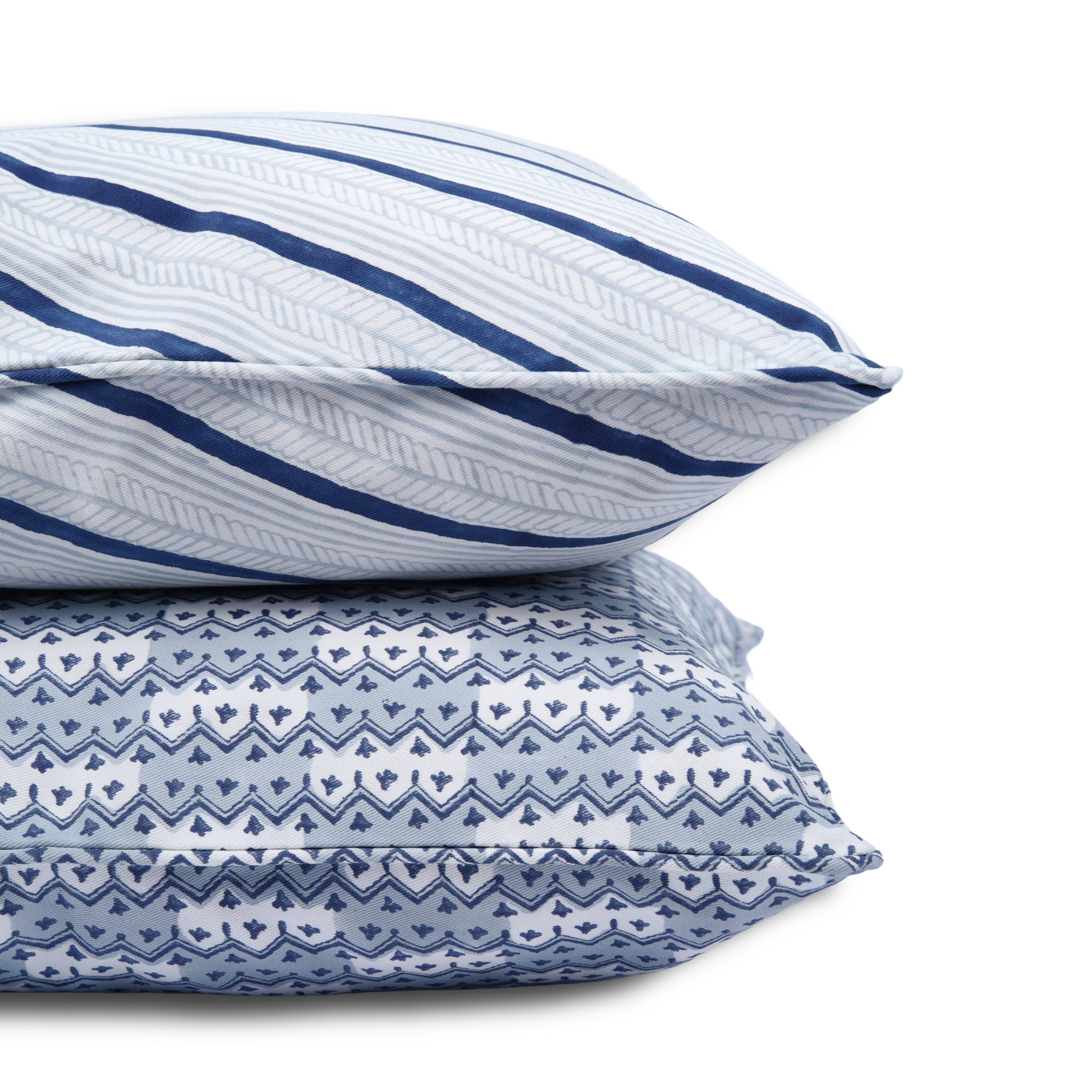 A close-up of two stacked hand-block printed cushions on a white background. The top pillow features Serere Frost indigo and light blue diagonal stripes, resting on a blue and white Munnar Frost cushion with delicate zig-zags. Piped edges highlight these handcrafted textiles, perfectly adding a cozy modern rustic Canadian farmhouse aesthetic to your home decor.