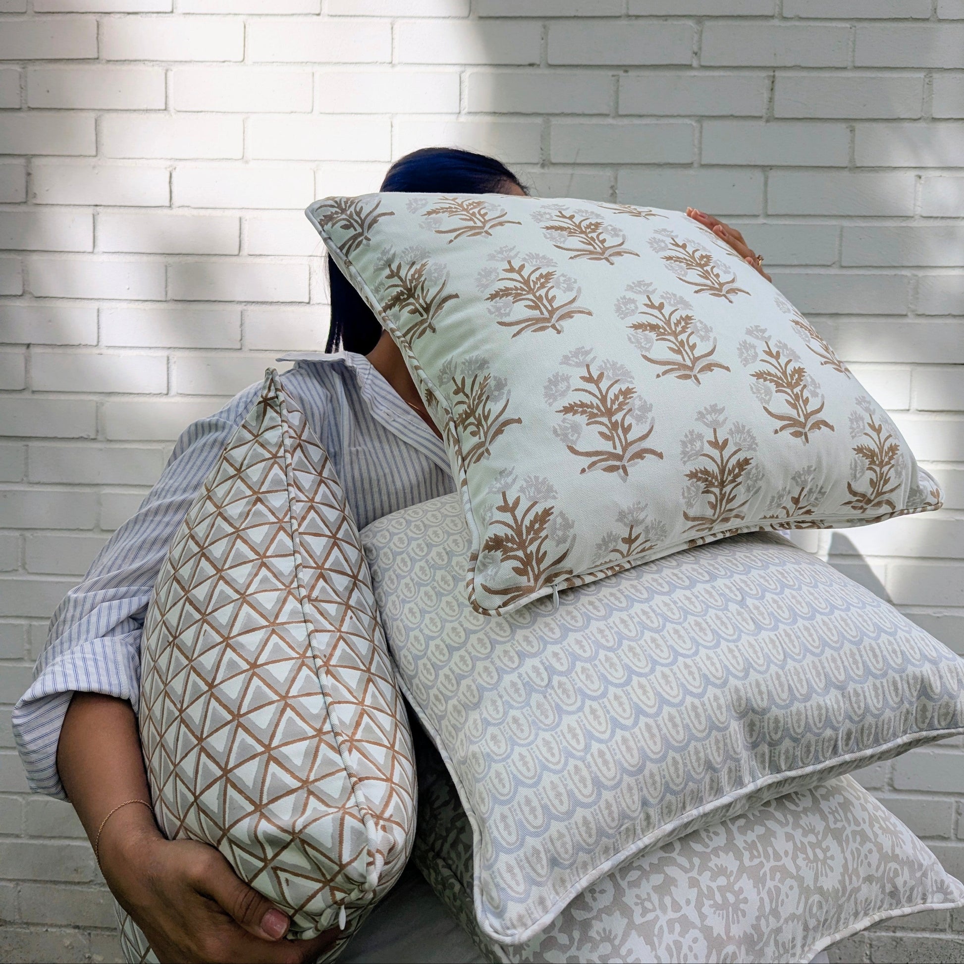 A lifestyle shot of a person holding a stack of four hand-block printed cushions outdoors against a white brick wall in dappled sunlight. The beautifully curated collection features earthy neutral tones, showcasing a tan botanical floral motif, a soft grey geometric scalloped arch pattern, and a tan geometric triangle design. These coordinating handcrafted textiles perfectly embody a cozy modern rustic Canadian farmhouse aesthetic for your home decor.