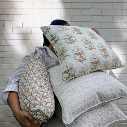 A lifestyle shot of a person holding a stack of four hand-block printed cushions outdoors against a white brick wall in dappled sunlight. The beautifully curated collection features earthy neutral tones, showcasing a tan botanical floral motif, a soft grey geometric scalloped arch pattern, and a tan geometric triangle design. These coordinating handcrafted textiles perfectly embody a cozy modern rustic Canadian farmhouse aesthetic for your home decor.