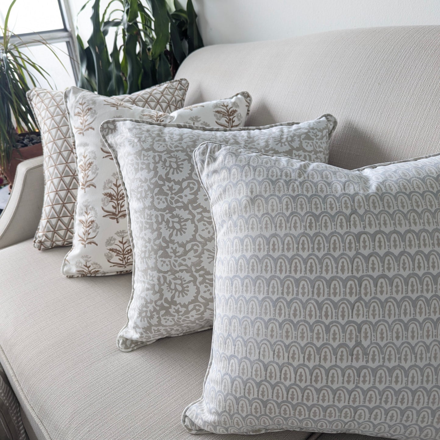 A beautiful arrangement of four hand-block printed cushions lined up on a neutral textured sofa. The coordinating collection features soft grey and earthy tan tones, showcasing a mix of geometric scalloped arches, elegant botanical florals, and triangle patterns. These elegantly handcrafted textiles perfectly add a cozy modern rustic Canadian farmhouse aesthetic to your living room decor.