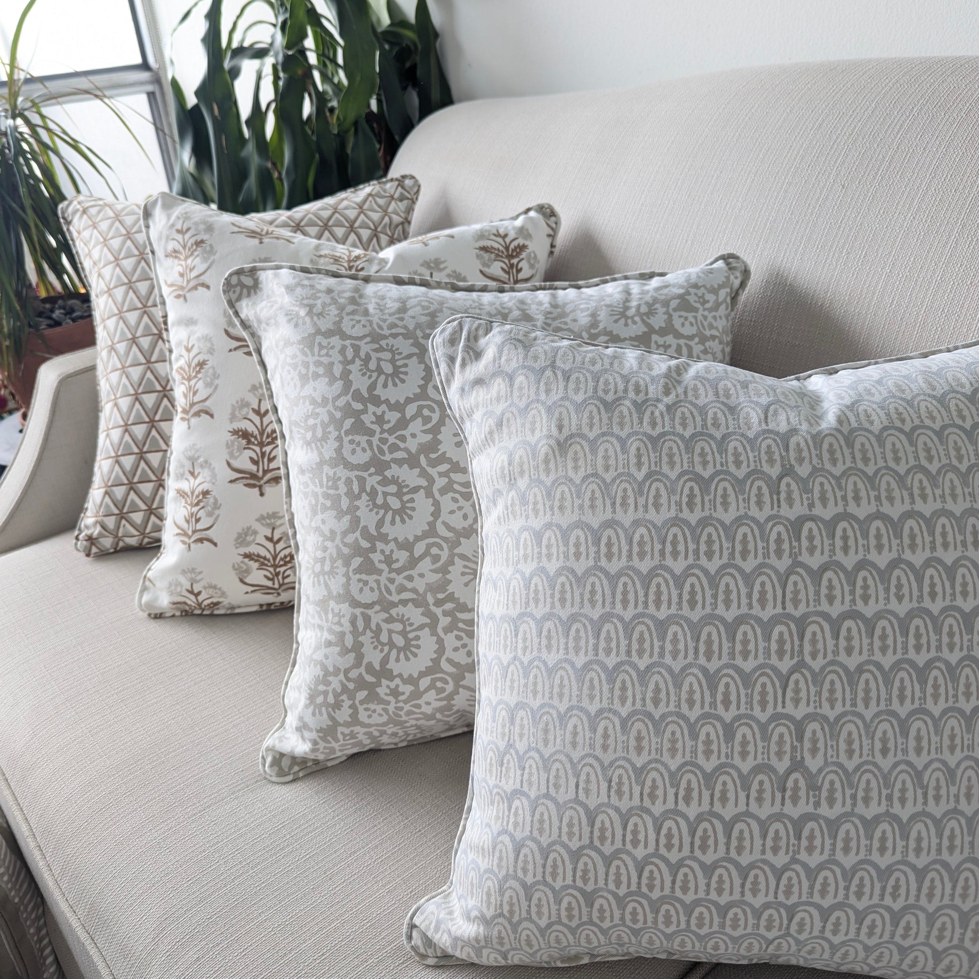 A beautiful arrangement of four hand-block printed cushions lined up on a neutral textured sofa. The coordinating collection features soft grey and earthy tan tones, showcasing a mix of geometric scalloped arches, elegant botanical florals, and triangle patterns. These elegantly handcrafted textiles perfectly add a cozy modern rustic Canadian farmhouse aesthetic to your living room decor.