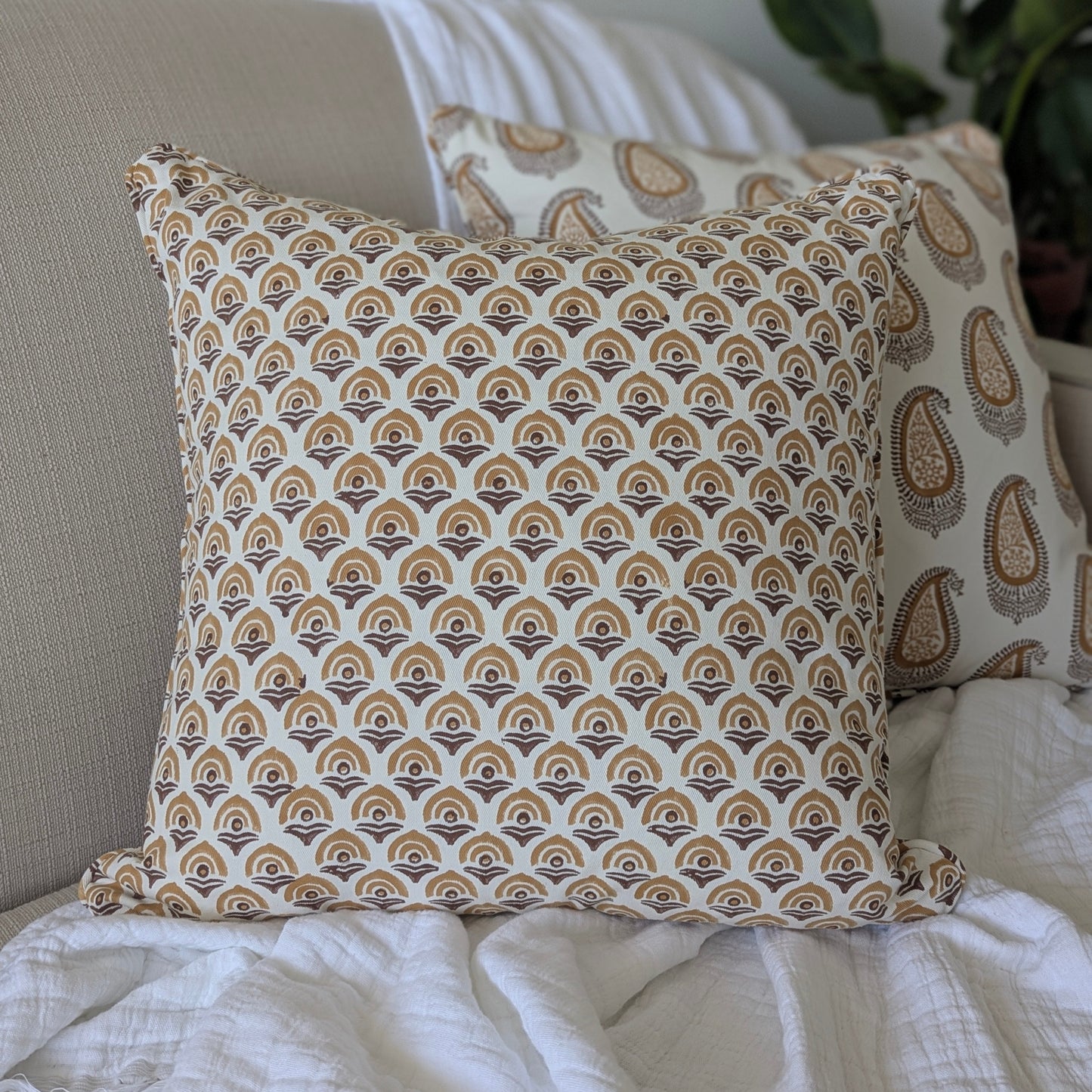 A close-up of a hand-block printed square cushion featuring a warm ochre and earthy brown geometric arch pattern on a cream background. Resting on a neutral beige sofa with a white textured throw, this handcrafted textile perfectly adds a cozy modern rustic Canadian farmhouse aesthetic to your living room decor.