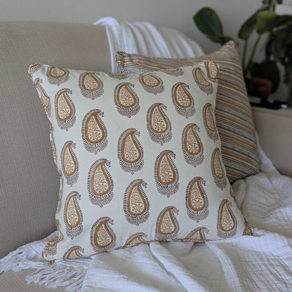 A close-up of a hand-block printed square cushion featuring an earthy brown and warm ochre paisley pattern on a cream background. Resting on a neutral textured sofa, this handcrafted textile perfectly adds a cozy modern rustic Canadian farmhouse aesthetic to your living room decor.