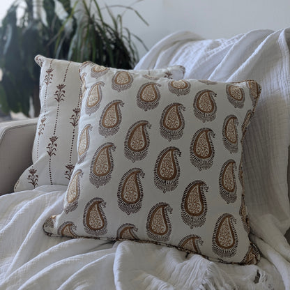 A close-up of a hand-block printed square cushion featuring an earthy brown and warm ochre paisley pattern, resting on a neutral sofa alongside a coordinating floral-striped pillow and a soft white throw blanket. These handcrafted textiles perfectly add a cozy modern rustic Canadian farmhouse aesthetic to your living room decor.