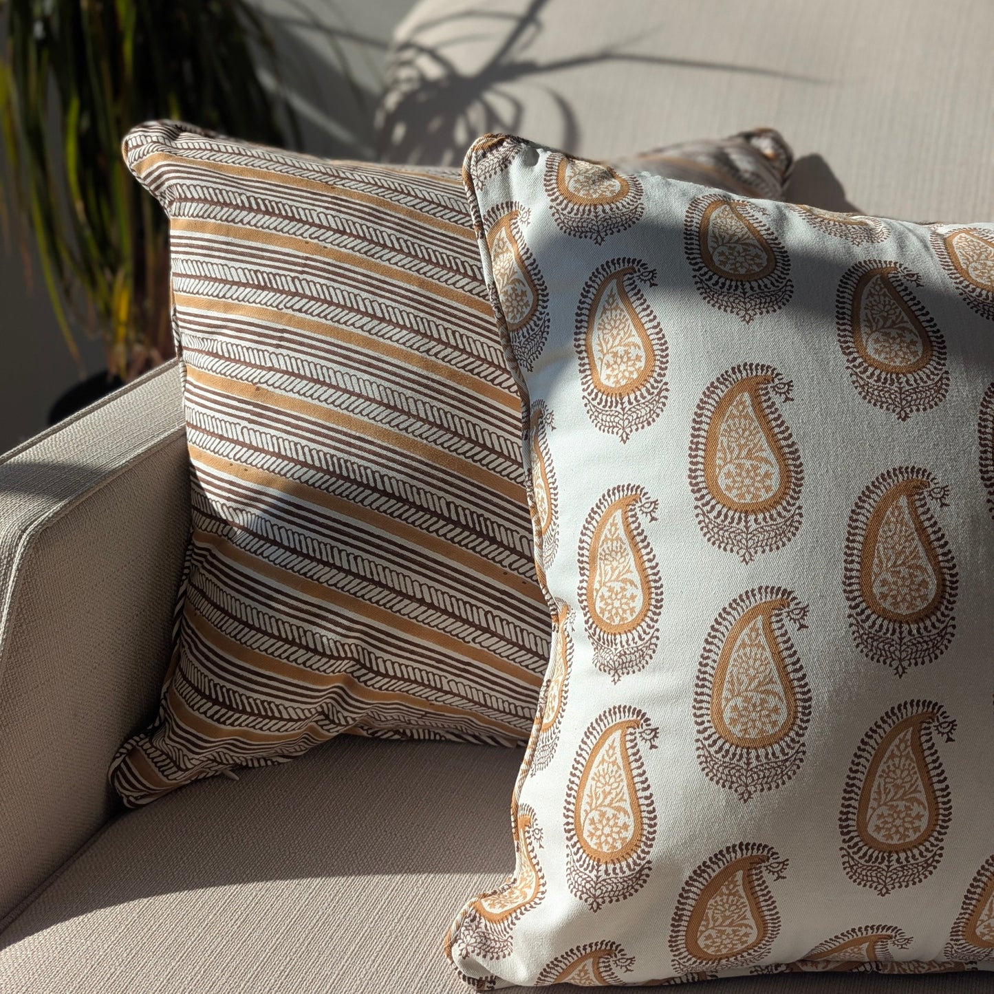 A close-up of two hand-block printed cushions resting on a neutral textured sofa, bathed in bright, natural sunlight. The foreground features a warm ochre and earthy brown classic paisley pattern, elegantly layered in front of a coordinating diagonal striped cushion. These beautifully handcrafted textiles perfectly add a cozy modern rustic Canadian farmhouse aesthetic to your living room decor.