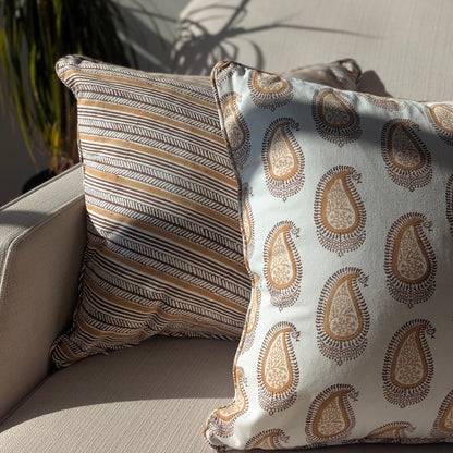 A close-up of two hand-block printed cushions resting on a neutral textured sofa, bathed in bright, natural sunlight. The foreground features a warm ochre and earthy brown classic paisley pattern, elegantly layered in front of a coordinating diagonal striped cushion. These beautifully handcrafted textiles perfectly add a cozy modern rustic Canadian farmhouse aesthetic to your living room decor.