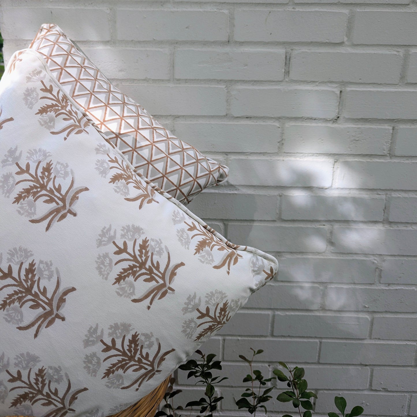 Two hand-block printed cushion covers resting against a white brick wall outdoors. The foreground pillow features an earthy brown floral sprig design, backed by a geometric triangle pattern. These artisanal textiles add a modern rustic farmhouse touch to Canadian and USA homes.