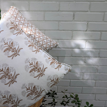 Two hand-block printed cushion covers resting against a white brick wall outdoors. The foreground pillow features an earthy brown floral sprig design, backed by a geometric triangle pattern. These artisanal textiles add a modern rustic farmhouse touch to Canadian and USA homes.