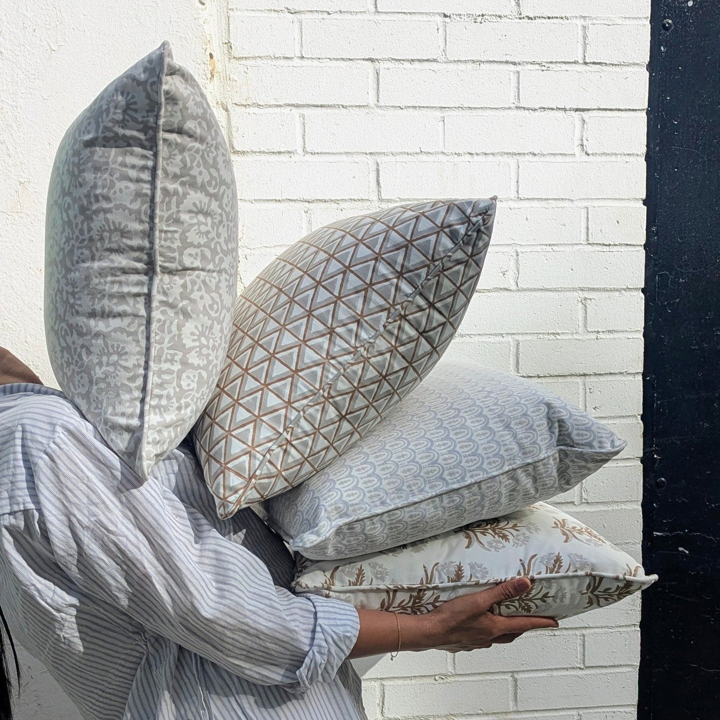 A lifestyle shot of a person carrying a beautifully curated stack of four hand-block printed cushions outdoors against a textured white brick wall in bright sunlight. The collection features earthy tan and soft grey tones, showcasing a mix of elegant botanical florals, geometric triangles, and scalloped arch patterns. These coordinating handcrafted textiles perfectly embody a cozy modern rustic Canadian farmhouse aesthetic for your home decor.