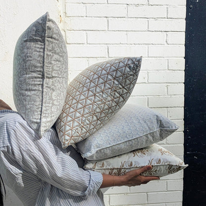 A lifestyle shot of a person carrying a beautifully curated stack of four hand-block printed cushions outdoors against a textured white brick wall in bright sunlight. The collection features earthy tan and soft grey tones, showcasing a mix of elegant botanical florals, geometric triangles, and scalloped arch patterns. These coordinating handcrafted textiles perfectly embody a cozy modern rustic Canadian farmhouse aesthetic for your home decor.