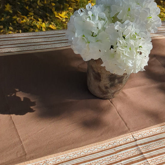 A brown tablecloth with a matching striped runner and a white floral centerpiece in a rustic vase, set outdoors on a grassy lawn with trees in the background.