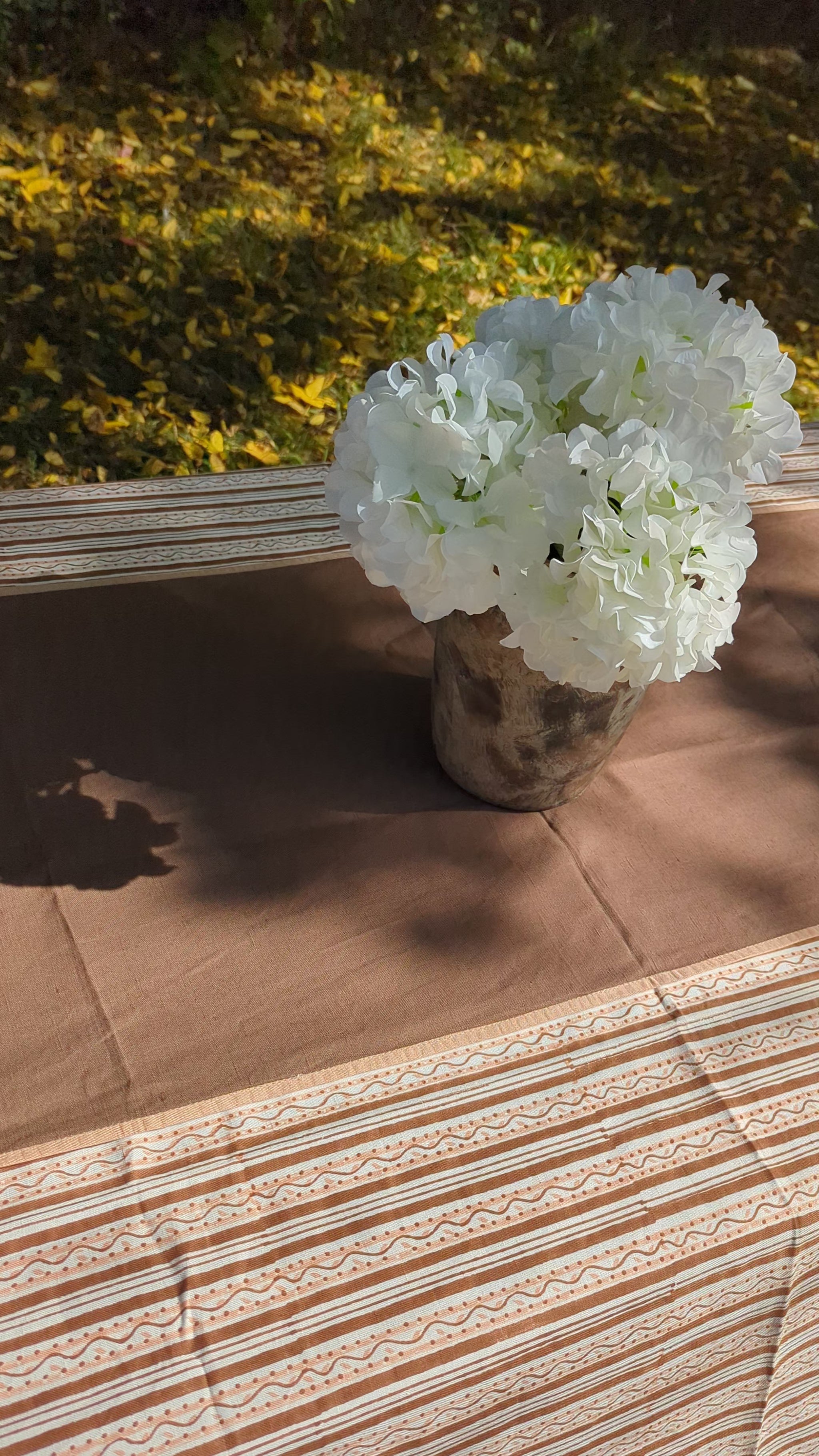 A brown tablecloth with a matching striped runner and a white floral centerpiece in a rustic vase, set outdoors on a grassy lawn with trees in the background.