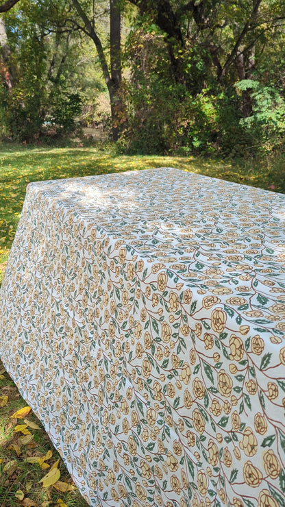 A rectangular outdoor table set with a cream tablecloth featuring a beige and green floral pattern, a matching scalloped runner, white plates, and folded dark green napkins. The background shows blurred green grass and foliage.