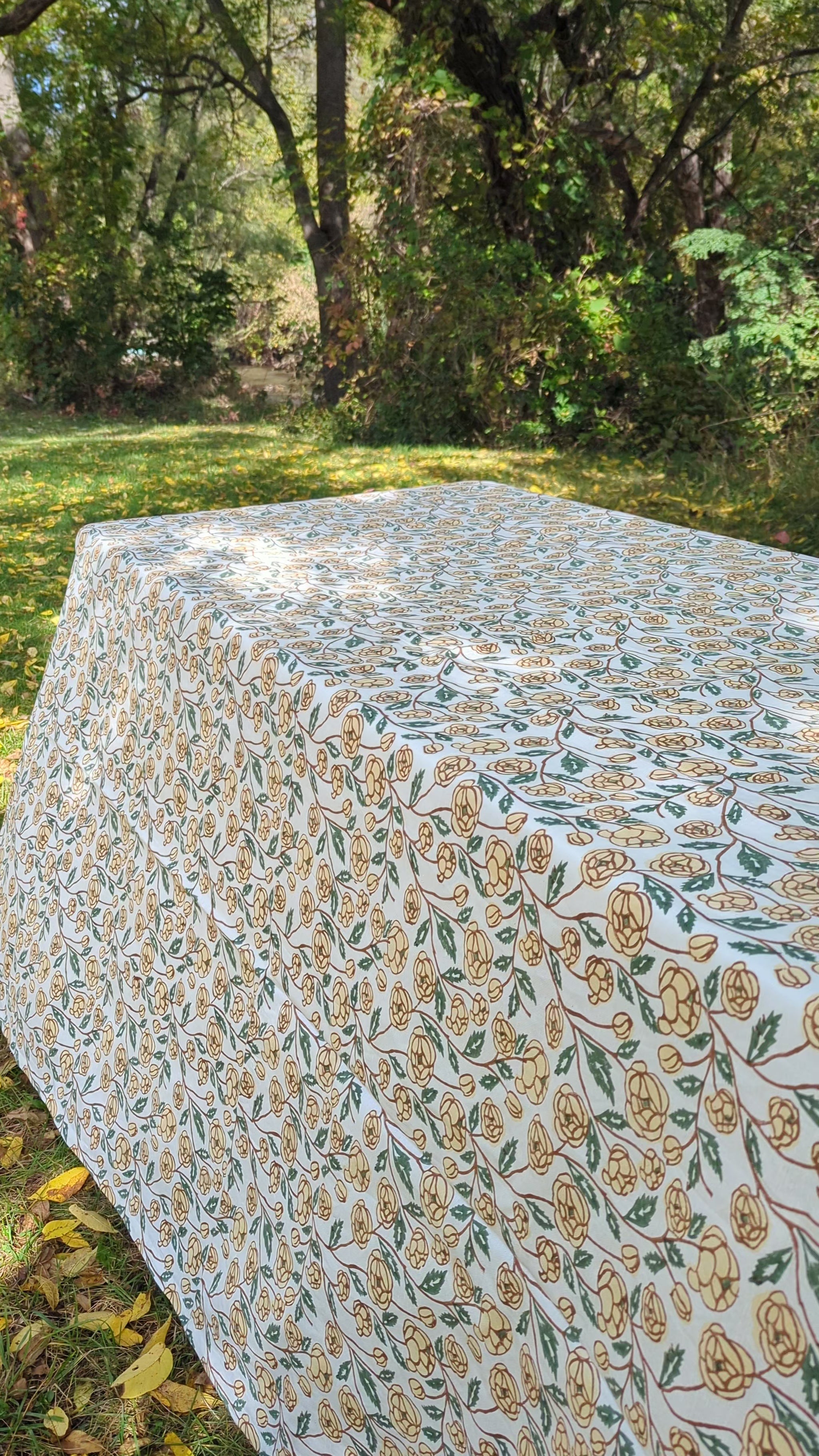 A rectangular outdoor table set with a cream tablecloth featuring a beige and green floral pattern, a matching scalloped runner, white plates, and folded dark green napkins. The background shows blurred green grass and foliage.