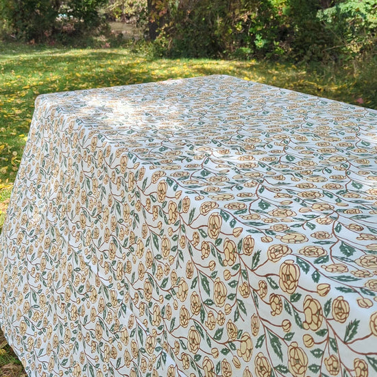 A top-down view of a table set with a beige tablecloth featuring a decorative floral border, two beige placemats, white plates, and dark green napkins with gold embroidered details.