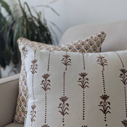 Close-up angle of the Reed Soil earthy brown floral 100% cotton block printed pillow cover layered in front of a coordinating brown scalloped pattern cushion on a light beige sofa. The soft-focus lifestyle shot features a blurred houseplant in the background, drawing attention to the crisp white fabric and intricate artisan hand-stamped textile details. Fine handmade home decor.