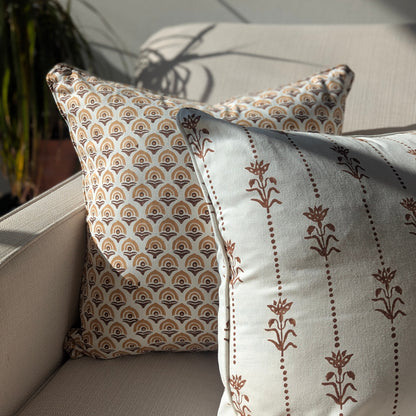 Close-up of the Reed Soil earthy brown floral 100% cotton block printed pillow cover paired with a coordinating brown scalloped pattern cushion on a light beige sofa. The cozy, sunlit scene features dramatic, artistic shadows from a nearby houseplant draped across the crisp white fabric, highlighting the fine handmade artisan textile home decor.