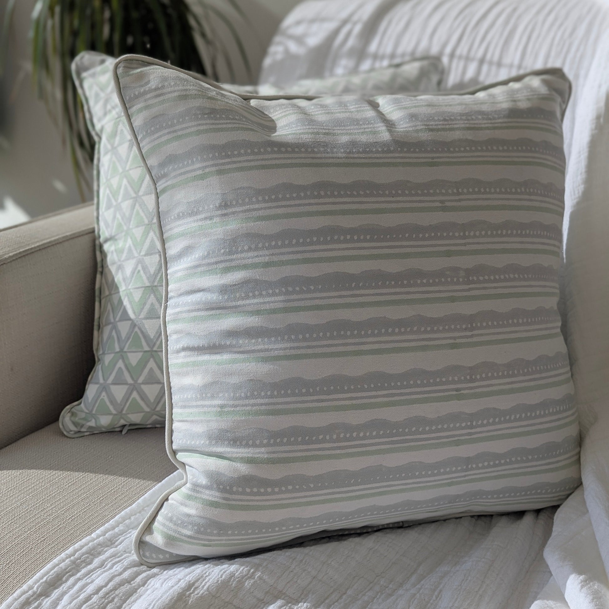 A close-up view of two hand-block printed cushions resting on a neutral textured sofa. The foreground features a cushion with a refreshing sage green and light blue horizontal striped and wavy pattern, elegantly layered in front of a coordinating geometric triangle pattern cushion. These beautifully handcrafted textiles perfectly add a cozy modern rustic Canadian farmhouse aesthetic to your living room decor.