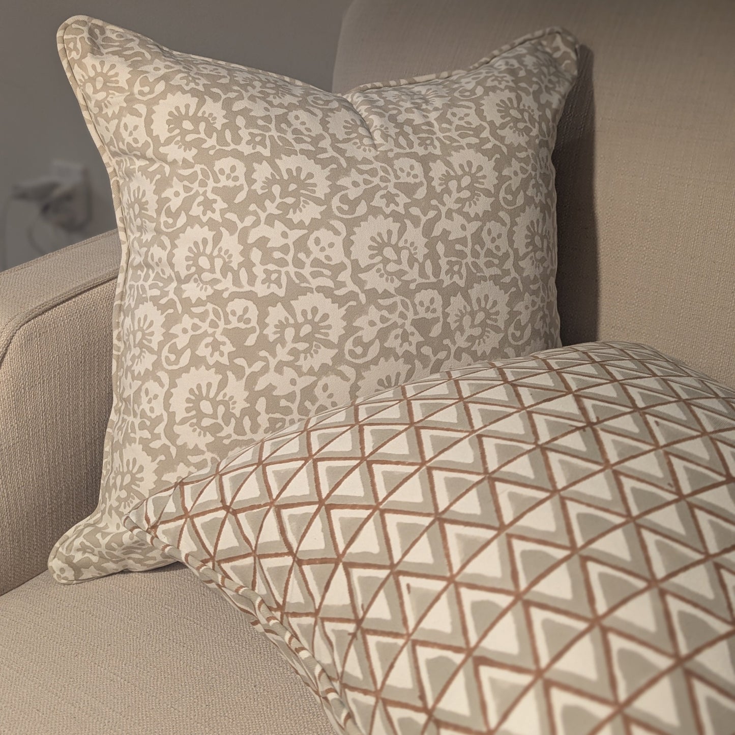 Two hand-block printed cushions resting on a cozy neutral sofa. The background cushion features an elegant soft grey botanical floral pattern, while the foreground cushion showcases a coordinating earthy tan and soft grey geometric triangle design. These elegantly handcrafted textiles perfectly add a modern rustic Canadian farmhouse aesthetic to your living room decor.