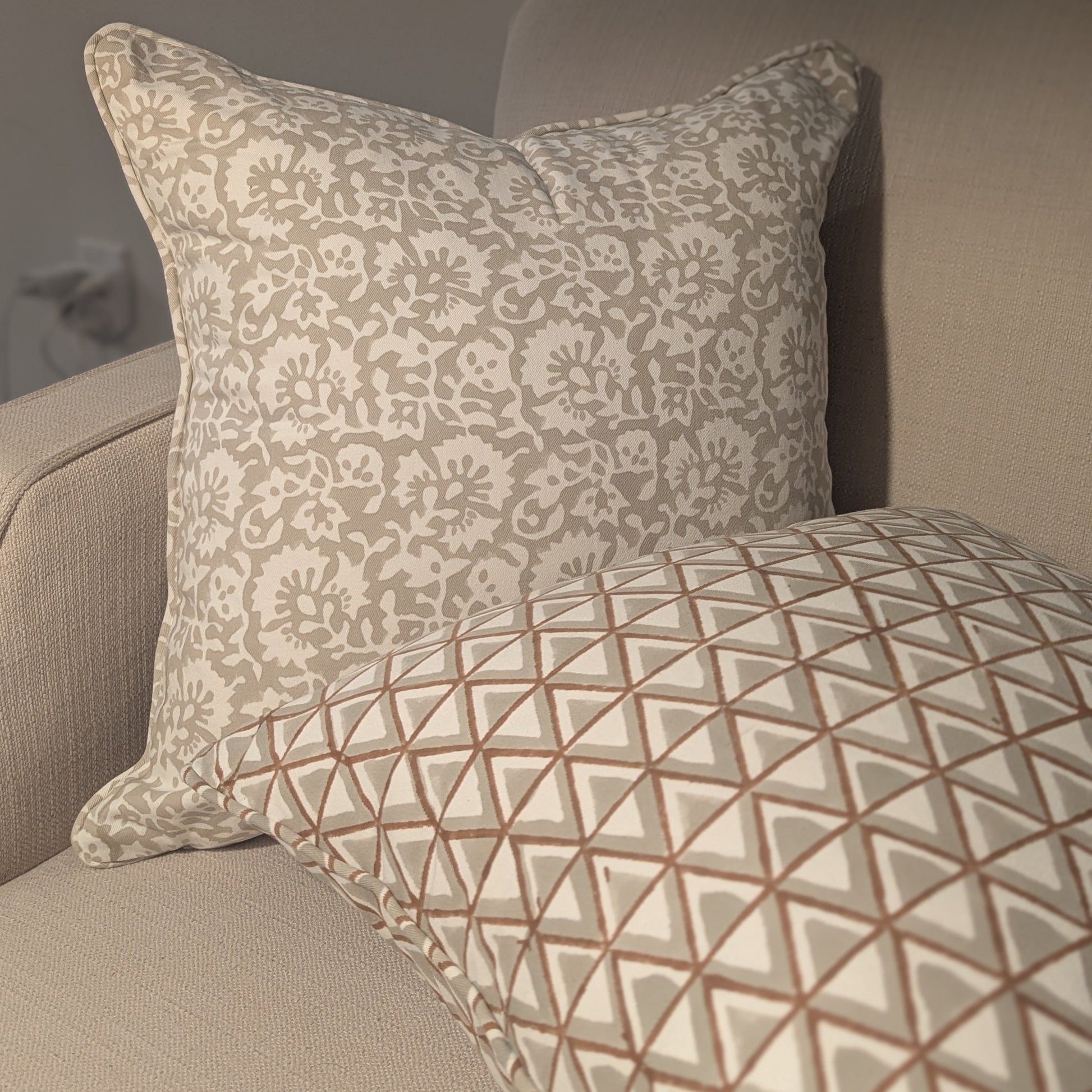 Two hand-block printed cushions resting on a cozy neutral sofa. The background cushion features an elegant soft grey botanical floral pattern, while the foreground cushion showcases a coordinating earthy tan and soft grey geometric triangle design. These elegantly handcrafted textiles perfectly add a modern rustic Canadian farmhouse aesthetic to your living room decor.