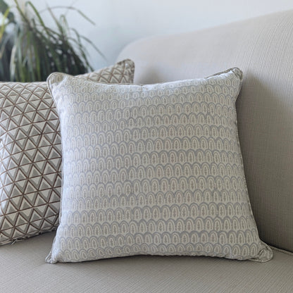 A close-up of a hand-block printed square cushion featuring a delicate, soft grey geometric scalloped arch pattern on a cream background. Resting on a neutral textured sofa alongside a coordinating patterned pillow, this handcrafted textile perfectly adds a cozy modern rustic Canadian farmhouse aesthetic to your living room decor.