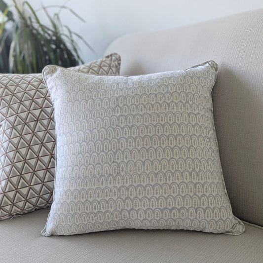 A close-up of a hand-block printed square cushion featuring a delicate, soft grey geometric scalloped arch pattern on a cream background. Resting on a neutral textured sofa alongside a coordinating patterned pillow, this handcrafted textile perfectly adds a cozy modern rustic Canadian farmhouse aesthetic to your living room decor.
