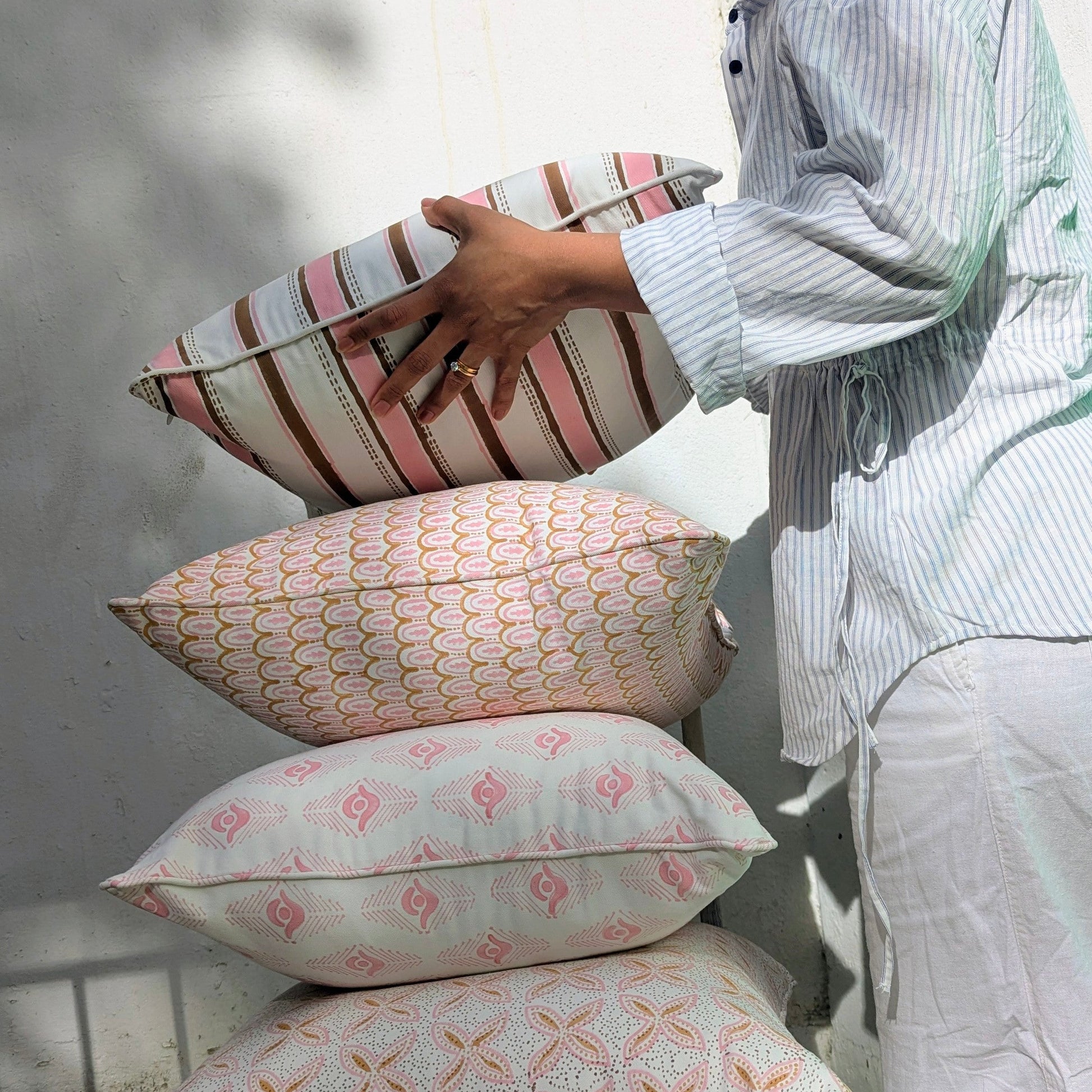 A person arranging a stack of four 100% cotton block printed throw pillow and cushion covers against a sunlit white wall. The stack features hand-printed pink, tan, and brown patterns including stripes, scalloped arches, geometric diamonds, and floral motifs. Soft handmade textile home decor collection.