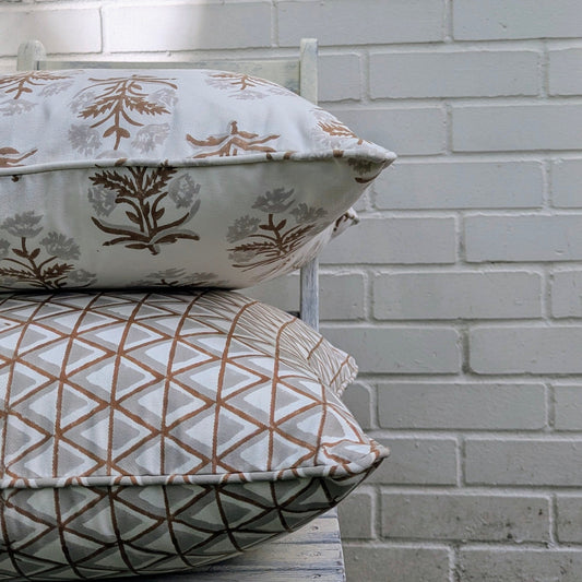 Two stacked hand-block printed cushion covers on a chair against a white brick wall. The top features an earthy brown floral sprig, while the bottom displays a geometric triangle pattern. With piped edges, this artisanal bundle brings modern rustic farmhouse charm to Canadian and USA homes.