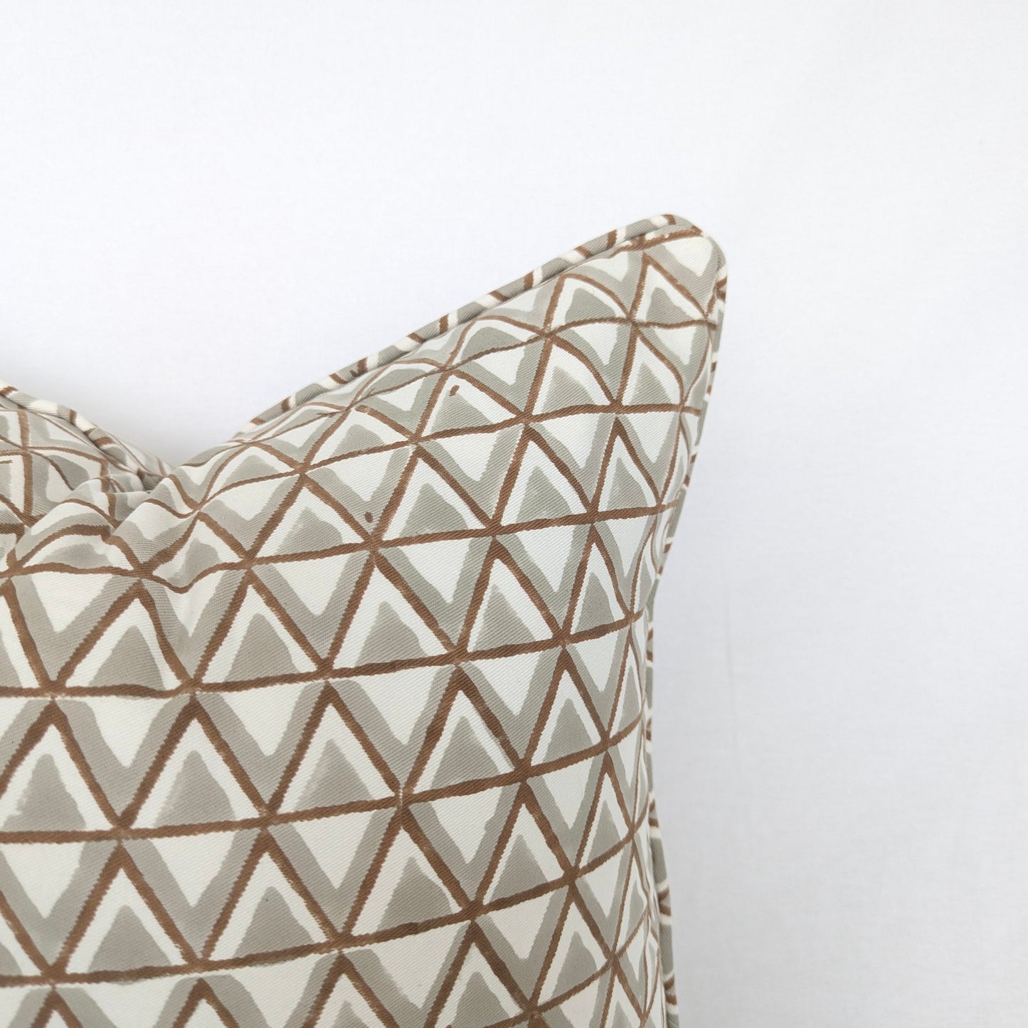A close-up view of the top corner of a hand-block printed cushion featuring an earthy tan and soft grey geometric triangle pattern on a cream background. This detail shot highlights the neatly finished piped edge, showcasing the quality of this handcrafted textile that perfectly adds a cozy modern rustic Canadian farmhouse aesthetic to any home decor.