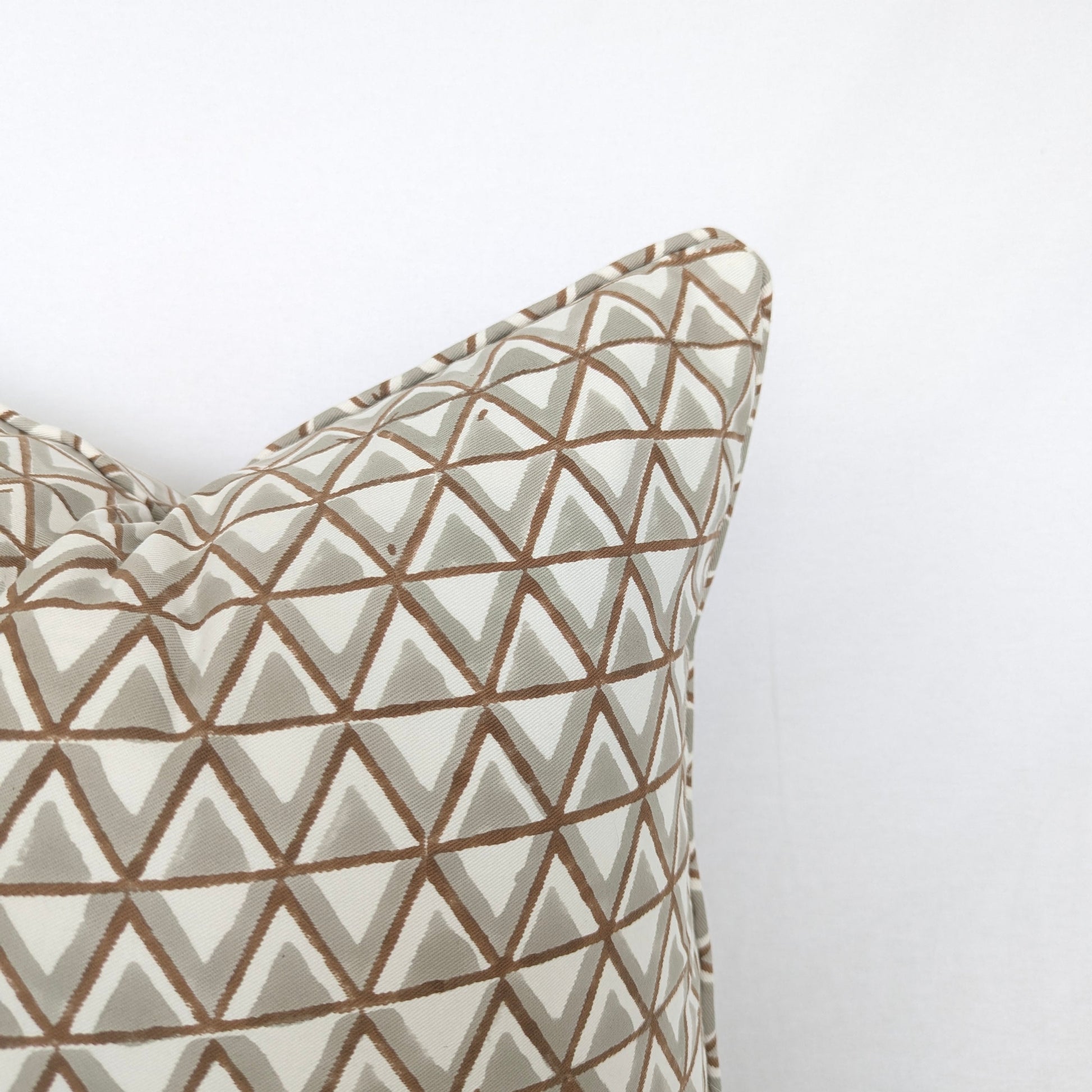 A close-up view of the top corner of a hand-block printed cushion featuring an earthy tan and soft grey geometric triangle pattern on a cream background. This detail shot highlights the neatly finished piped edge, showcasing the quality of this handcrafted textile that perfectly adds a cozy modern rustic Canadian farmhouse aesthetic to any home decor.